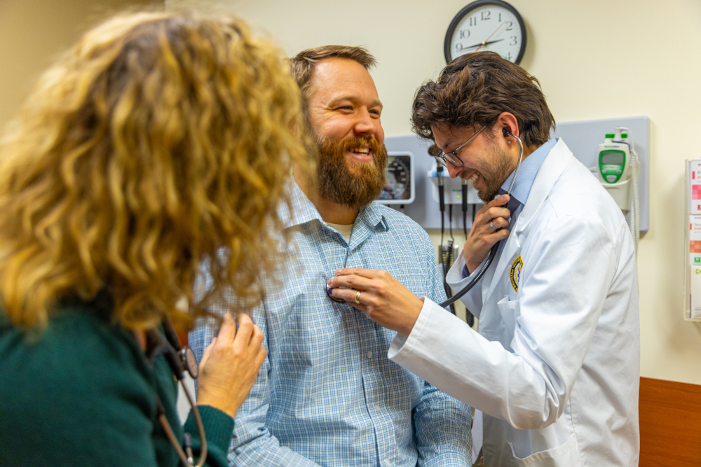 A Physician Assistant student in a white lab coat laughs while listening to a patient's heart with a stethoscope