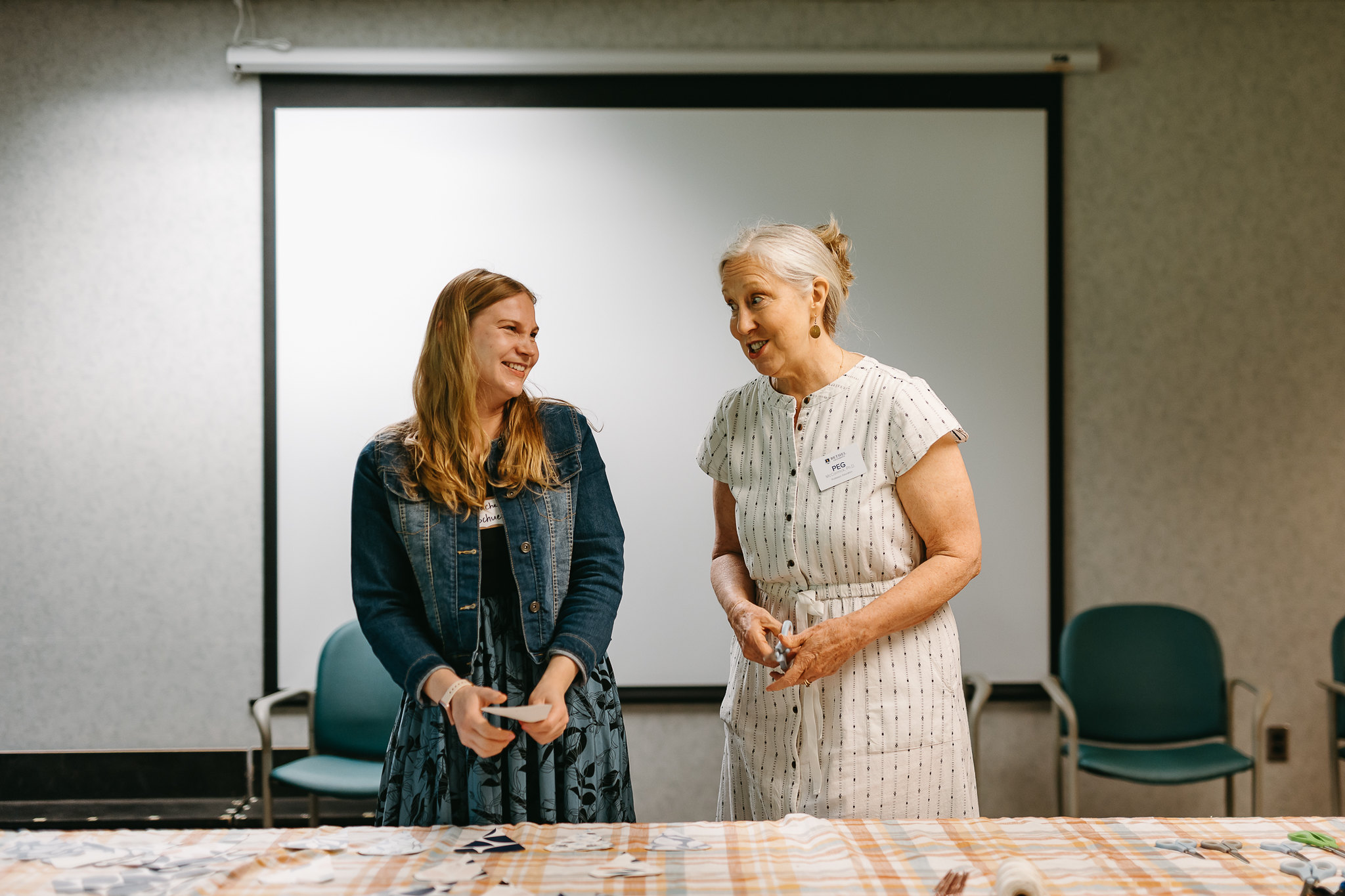 Two women smile and talk while leading a hands-on activity at a table in front of a projection screen.