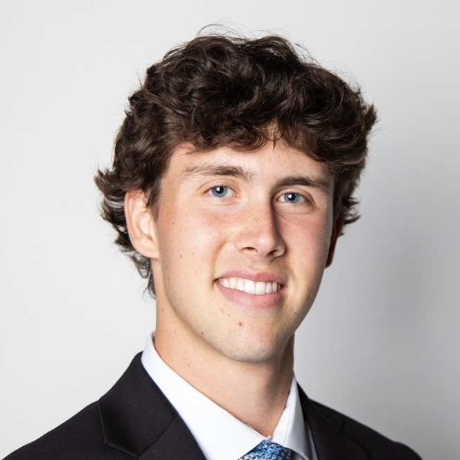 Jacob Merriam wears a dark suit and blue patterned tie for a professional headshot.